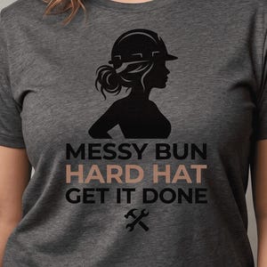 Peut inclure: T-shirt gris foncé avec une silhouette noire d'une femme portant un casque de chantier et un chignon décoiffé. Le texte "MESSY BUN HARD HAT GET IT DONE" est imprimé en dessous, avec un marteau et une clé croisés.