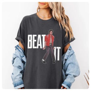 May include: Dark gray t-shirt featuring the text "BEAT IT" in white block letters, with an image of a person in a red jacket. The shirt has a relaxed fit and short sleeves. The overall style is casual and vintage.
