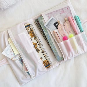 May include: A white fabric organizer case containing two spiral-bound notebooks, a clear plastic ruler, a small photo, two wing-shaped hair clips, a glittery pen, two highlighters, and two markers. The case features a zippered pocket.