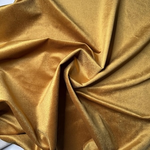 Può includere: Primo piano di un tessuto di velluto giallo dorato con morbide pieghe e una lucentezza lucida. Il tessuto è drappeggiato, creando ombre e riflessi che ne esaltano la consistenza. Il materiale appare spesso e lussuoso.
