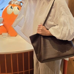May include: A dark brown tote bag with a textured surface. The bag has a long shoulder strap and is being held by a person wearing a white blouse. An orange cartoon figure is in the background.