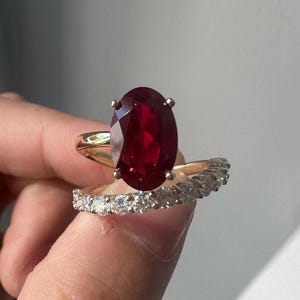 以下が含まれることがあります： 4本の爪で留められた大きなオーバルカットの深い赤色の宝石が特徴のゴールドリングセット。磨き上げられたイエローゴールドの台座に、小さな丸い透明なホワイトダイヤモンドが一列に並んだ、お揃いのバンドリングが組み合わされています。