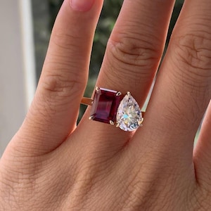 Può includere: Un anello in oro con due pietre prominenti affiancate: una gemma rettangolare rosso scuro con taglio a smeraldo e una gemma trasparente a forma di goccia. Le pietre sono fissate da griffe in oro su un sottile anello d'oro lucido.