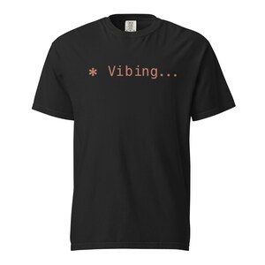 Op de afbeelding: Zwart T-shirt met de tekst "* Vibing..." in koraal. Het shirt met korte mouwen en ronde hals is gemaakt van een zacht materiaal. De tekst staat in het midden op de voorkant.