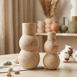 May include: Two beige travertine vases with a stacked, double-sphere silhouette and a porous, textured surface. The smaller vase features a small, square, light brown leather tag tied around its neck with a thin string, displaying a simple line drawing.