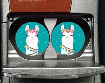 May include: Two turquoise blue car coasters with a white llama wearing a flower crown and colorful tassels. The llama has its eyes closed and a peaceful expression.