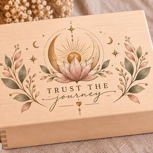 Könnte beinhalten: Eine rechteckige, helle Holzschatulle mit einem aufgedruckten Motiv aus einer Lotusblüte, einer Mondsichel und Sonnenstrahlen. Der Text "TRUST THE journey" ist mittig aufgedruckt. Die Box weist an den Ecken sichtbare Fingerzinken auf.
