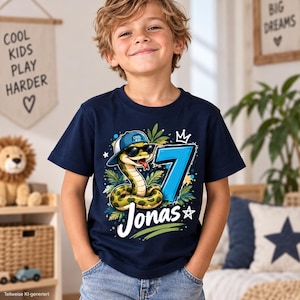 Könnte beinhalten: Ein dunkelblaues Kurzarm-T-Shirt mit einer Grafik einer grünen Schlange, die eine blau-weiße Baseballkappe und eine schwarze Sonnenbrille trägt. Das Motiv zeigt zudem eine große blaue Zahl 7, den Namen Jonas in weißer Schrift und Blätter.