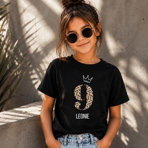 Könnte beinhalten: Ein schwarzes Kurzarm-T-Shirt mit einer großen Zahl 9 im Leopardenmuster, einer weißen, linienförmigen Krone über der Zahl und dem Namen LEONIE, der in weißen Großbuchstaben unter der Zahl aufgedruckt ist.