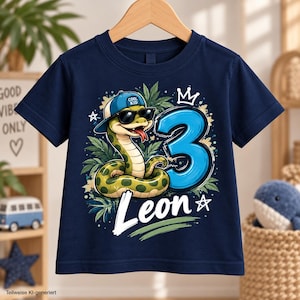 Könnte beinhalten: Ein dunkelblaues Kurzarm-T-Shirt mit einer Grafik einer grünen Schlange, die eine blaue Baseballkappe mit der Aufschrift COOL KID und eine schwarze Sonnenbrille trägt. Das Motiv zeigt eine große blaue 3, eine Krone und den Namen Leon in Weiß.
