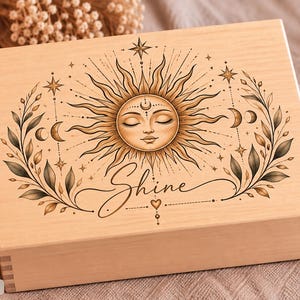 Könnte beinhalten: Eine rechteckige, helle Holzschatulle mit einer detaillierten Illustration einer Sonne mit menschlichem Gesicht, geschlossenen Augen und strahlenden Strahlen. Das Design umfasst Mondsicheln, Sterne und Blattzweige sowie das Wort Shine in Schreibschrift.