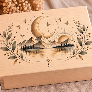 Könnte beinhalten: Eine rechteckige, helle Holzschatulle mit einer detaillierten schwarz-goldenen Illustration auf dem Deckel. Das Motiv zeigt eine Mondsichel, eine Sonne, Berggipfel, einen Wald und einen See, umrahmt von zarten Blattzweigen und kleinen Sternen.