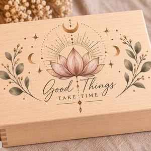Könnte beinhalten: Eine rechteckige, helle Holzschatulle mit einem Aufdruck, der eine rosa Lotusblüte, himmlische Mondphasen und belaubte Zweige zeigt. Der Text "Good Things TAKE TIME" ist mittig auf dem Deckel in einer schwarzen Schriftart abgebildet.