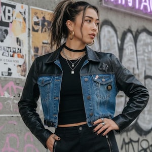May include: A cropped denim jacket with black leather sleeves and collar. The jacket has a blue denim body and black leather sleeves. The background features graffiti art and posters.