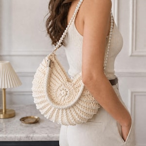 May include: Cream-colored, crocheted handbag with a pearl strap. The bag has a seashell-like design with a rounded shape and a textured surface. The strap is made of small, white pearls. The bag is worn over the shoulder.