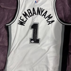 以下が含まれることがあります： 黒いトリムが施された白いバスケットボールジャージ。背中にはWEMBANYAMAの文字がアーチ状に配置され、その下に黒い数字の1がプリントされています。数字の1にはサインとPSA認証ステッカーが貼られています。首元にはNBAのロゴがあります。