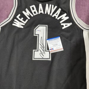 May include: A black basketball jersey featuring the name WEMBANYAMA and the number 1 in white lettering. The number 1 includes a black signature and a PSA authentication sticker. A PSA certificate of authenticity card is placed on the jersey.