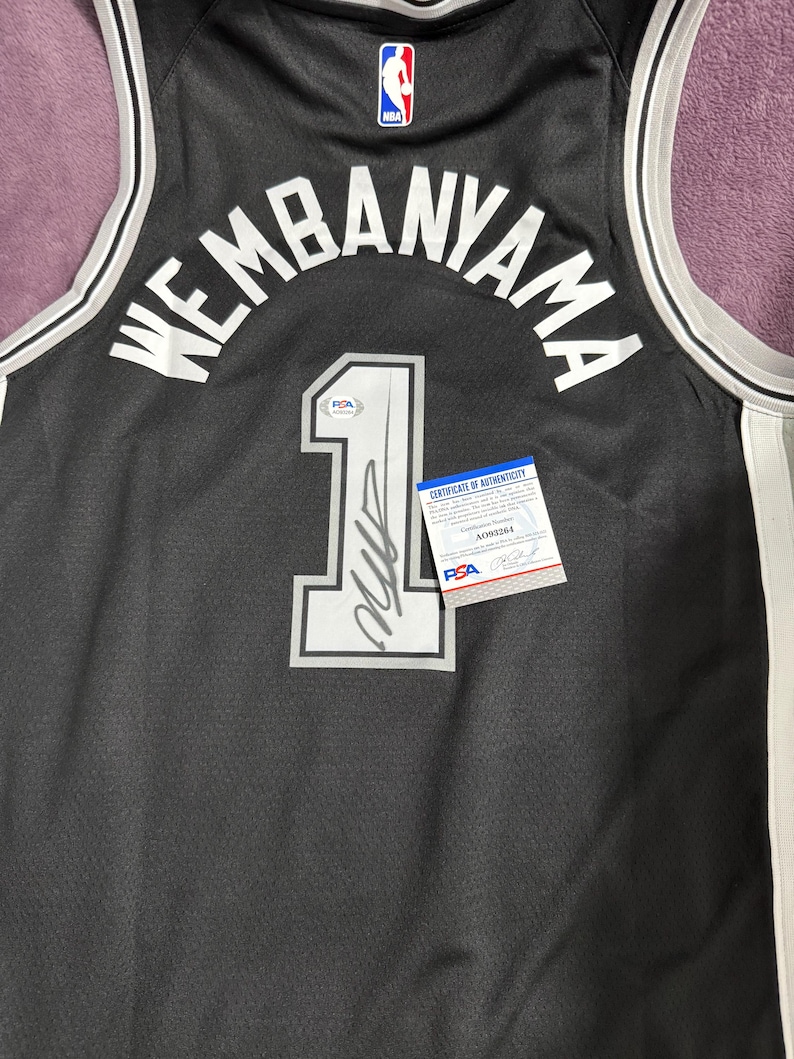 May include: A black basketball jersey featuring the name WEMBANYAMA and the number 1 in white lettering. The number 1 includes a black signature and a PSA authentication sticker. A PSA certificate of authenticity card is placed on the jersey.