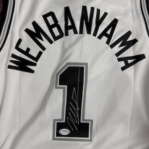 以下が含まれることがあります： 背面に黒い文字でWEMBANYAMAと記された白いバスケットボールジャージ。名前の下にはグレーの縁取りが施された黒い背番号1があり、サインとPSA認証の小さなステッカーが数字の上に貼られています。