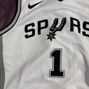 以下が含まれることがあります： 文字「U」が拍車のアイコンになった黒い「SPURS」の文字、グレーの縁取りがある黒い背番号「1」、黒いナイキのロゴ、Vネックとサイドパネルに黒とグレーのトリムがあしらわれた、白いバスケットボール用ジャージ。