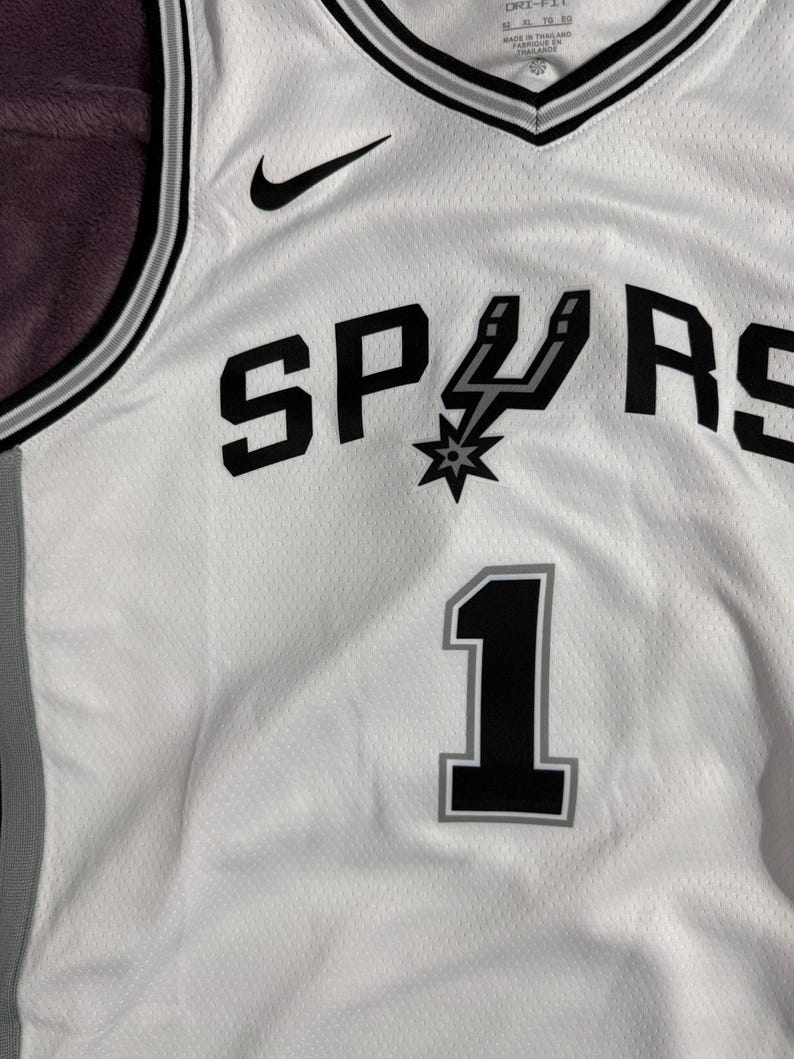 以下が含まれることがあります： 文字「U」が拍車のアイコンになった黒い「SPURS」の文字、グレーの縁取りがある黒い背番号「1」、黒いナイキのロゴ、Vネックとサイドパネルに黒とグレーのトリムがあしらわれた、白いバスケットボール用ジャージ。