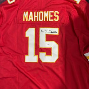 Puede incluir: Camiseta de f&uacute;tbol roja con el nombre "MAHOMES" y el n&uacute;mero "15" en blanco con ribete dorado. La camiseta tambi&eacute;n presenta los n&uacute;meros "5" y "1" en los hombros. La camiseta est&aacute; firmada.