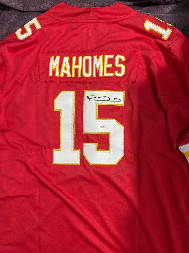 Puede incluir: Camiseta de f&uacute;tbol roja con el nombre "MAHOMES" y el n&uacute;mero "15" en blanco con ribete dorado. La camiseta tambi&eacute;n presenta los n&uacute;meros "5" y "1" en los hombros. La camiseta est&aacute; firmada.