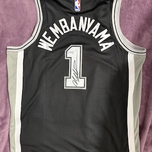 May include: A black basketball jersey featuring the name WEMBANYAMA and the number 1 in white lettering. The jersey includes a white and grey trim, an NBA logo at the neckline, and a signature on the number 1 with a PSA authentication sticker.
