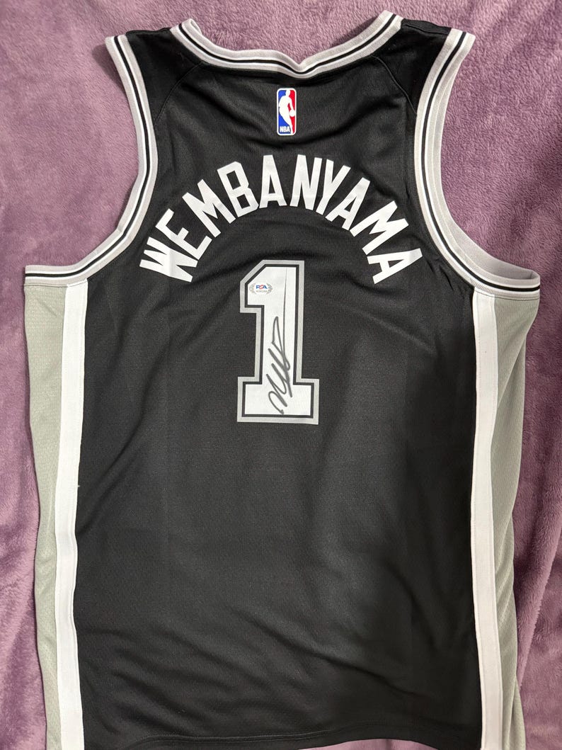 May include: A black basketball jersey featuring the name WEMBANYAMA and the number 1 in white lettering. The jersey includes a white and grey trim, an NBA logo at the neckline, and a signature on the number 1 with a PSA authentication sticker.