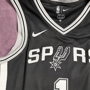 May include: A black basketball jersey featuring the San Antonio Spurs logo and the number 1 in white. The jersey has a V-neck with grey and white trim, a Nike swoosh logo, and Dri-Fit branding on the interior collar. The fabric has a mesh texture.