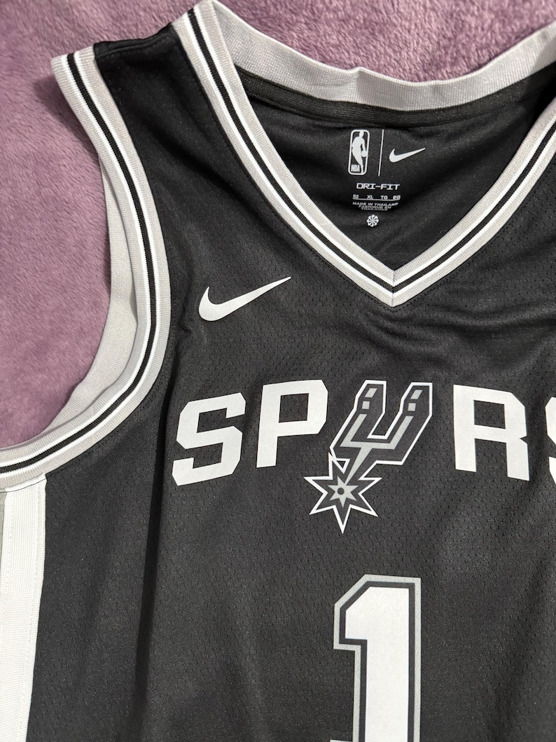 May include: A black basketball jersey featuring the San Antonio Spurs logo and the number 1 in white. The jersey has a V-neck with grey and white trim, a Nike swoosh logo, and Dri-Fit branding on the interior collar. The fabric has a mesh texture.
