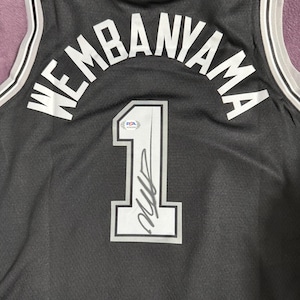May include: A black basketball jersey featuring the name WEMBANYAMA and the number 1 in white lettering with grey outlines. The jersey includes an NBA logo at the top and a PSA authentication sticker placed on the number 1, which also features a signature.