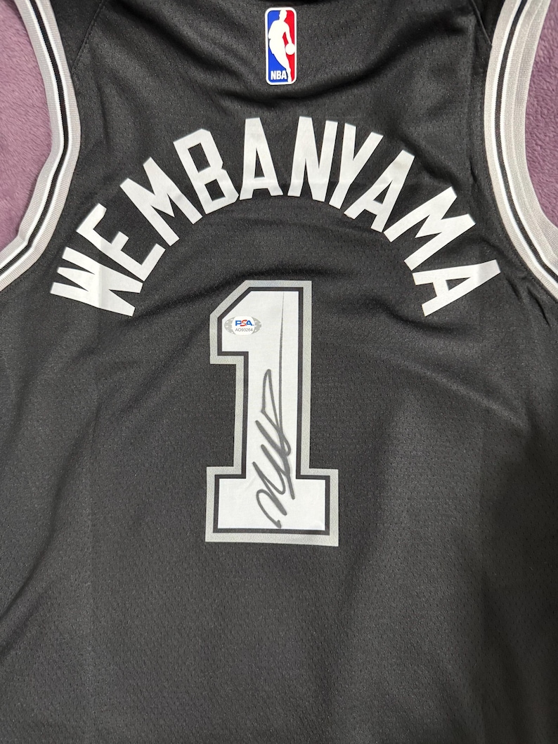 May include: A black basketball jersey featuring the name WEMBANYAMA and the number 1 in white lettering with grey outlines. The jersey includes an NBA logo at the top and a PSA authentication sticker placed on the number 1, which also features a signature.