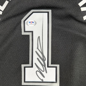 May include: A black mesh sports jersey featuring a large white number 1 with a grey and black outline. The number is signed in black ink and includes a PSA authentication sticker with the code AO93264. Partial white text is visible at the top.