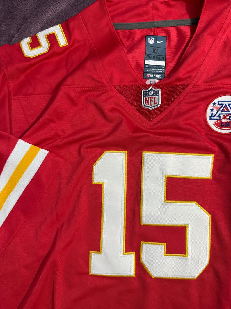 Puede incluir: Una camiseta de f&uacute;tbol americano de la NFL roja con el n&uacute;mero 15 en blanco con ribete amarillo. La camiseta tiene rayas blancas y amarillas en las mangas y un logotipo de la NFL en el pecho. La camiseta es talla XL.