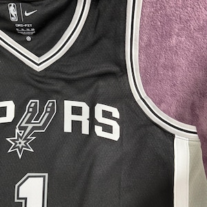 May include: A black basketball jersey featuring white trim on the V-neck and armholes. The front displays the San Antonio Spurs logo, the text "SPURS", and the number "1" in white. It includes a Nike swoosh, NBA logo, and Dri-Fit branding.