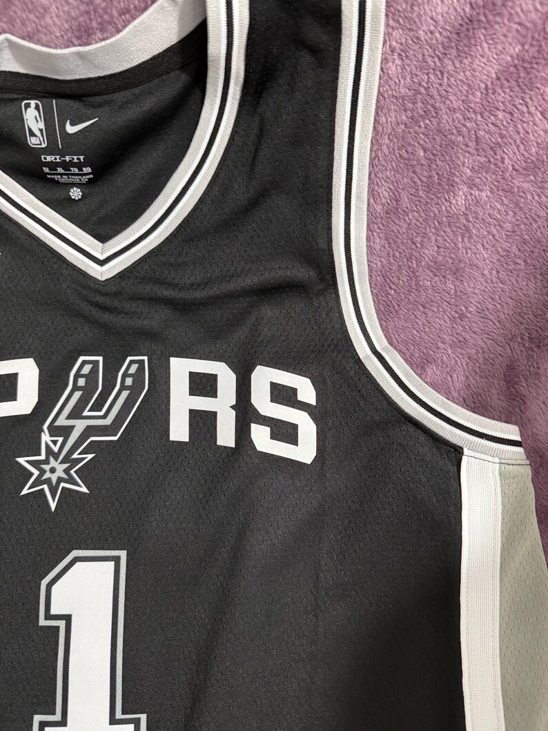 May include: A black basketball jersey featuring white trim on the V-neck and armholes. The front displays the San Antonio Spurs logo, the text "SPURS", and the number "1" in white. It includes a Nike swoosh, NBA logo, and Dri-Fit branding.