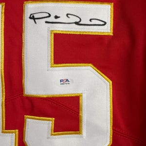 Puede incluir: Primer plano de una camiseta roja con un gran n&uacute;mero 5 blanco con contorno dorado. El n&uacute;mero est&aacute; cosido a la camiseta. La camiseta tiene un parche blanco con una firma y una pegatina de autenticaci&oacute;n PSA.