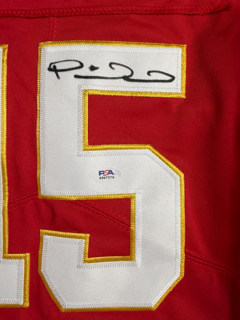 Puede incluir: Primer plano de una camiseta roja con un gran n&uacute;mero 5 blanco con contorno dorado. El n&uacute;mero est&aacute; cosido a la camiseta. La camiseta tiene un parche blanco con una firma y una pegatina de autenticaci&oacute;n PSA.
