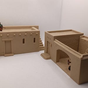 Könnte beinhalten: Zwei beigefarbene Miniaturgebäude, möglicherweise für ein Modell oder Diorama. Eines hat ein flaches Dach mit einer kleinen Figur darauf. Das andere hat einen gewölbten Eingang und einen offenen Innenraum. Die Gebäude haben eine strukturierte Oberfläche.