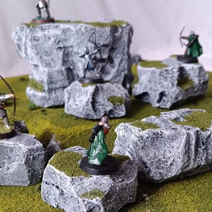May include: Miniature figurines of fantasy characters stand on gray, textured rock formations with patches of green moss. The rocks are set on a green, grassy surface, creating a diorama scene. The figurines are in various poses, some with bows and arrows.