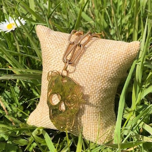 Peut inclure: Un pendentif en forme de crâne doré avec des motifs floraux, attaché à une chaîne. Le pendentif est présenté sur un petit coussin en toile de jute beige, sur fond d'herbe verte et d'une marguerite.