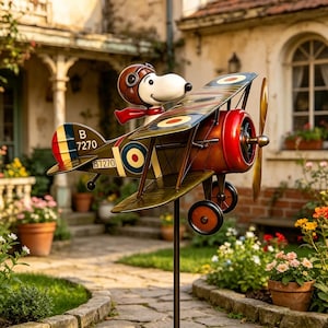 Può includere: Un paletto da giardino in metallo con Snoopy su un aereo biplano. L'aereo è dipinto in verde oliva, rosso e marrone con un'elica e ruote. Snoopy indossa un casco da pilota e una sciarpa rossa. L'aereo ha il numero B 7270.