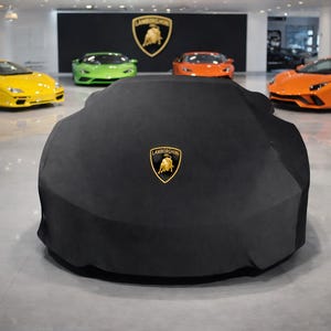 Puede incluir: Una funda negra para coche con el logotipo dorado de Lamborghini cubre un vehículo en una sala de exposición. Varios coches deportivos Lamborghini de colores son visibles en el fondo, incluyendo modelos amarillos, verdes y naranjas.