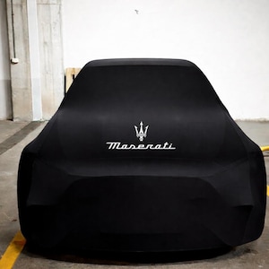 Puede incluir: Una funda negra para coche con el logotipo blanco de Maserati. La funda cubre un vehículo, ocultando su forma. El logotipo de Maserati, un tridente sobre el nombre de la marca, es visible. El coche está aparcado en un garaje.