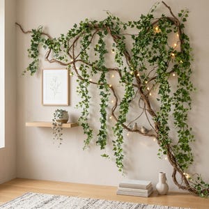 Puede incluir: Una decoración mural con falsas enredaderas de hiedra que caen de una estructura de ramas marrones. La exhibición incluye una impresión botánica enmarcada, una pequeña estantería con una planta en maceta y luces de cadena. Una alfombra y un jarrón están en primer plano.