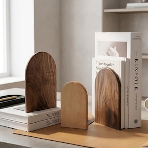Puede incluir: Juego de sujetalibros de madera de estilo minimalista. Dos son de color marrón oscuro y uno de color marrón claro. Los sujetalibros tienen una parte superior arqueada y sostienen libros con cubiertas blancas. Los libros están sobre una superficie de cuero marrón claro.