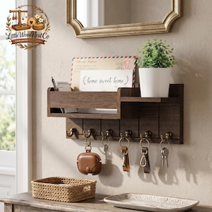 May include: A dark brown wooden wall-mounted organiser with a shelf, mail slot, and key hooks. The organiser has a small potted plant, a notepad, and a card that says "home sweet home." A small woven basket and a decorative tray are on the table below.