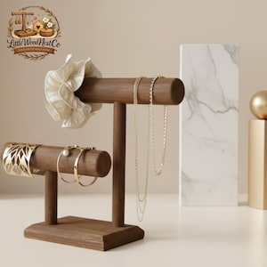 Puede incluir: Un expositor de joyas de madera marrón oscuro. Exhibe una variedad de joyas, incluyendo collares de oro, pulseras y una goma para el pelo blanca. El expositor tiene una base rectangular y un diseño en forma de T.