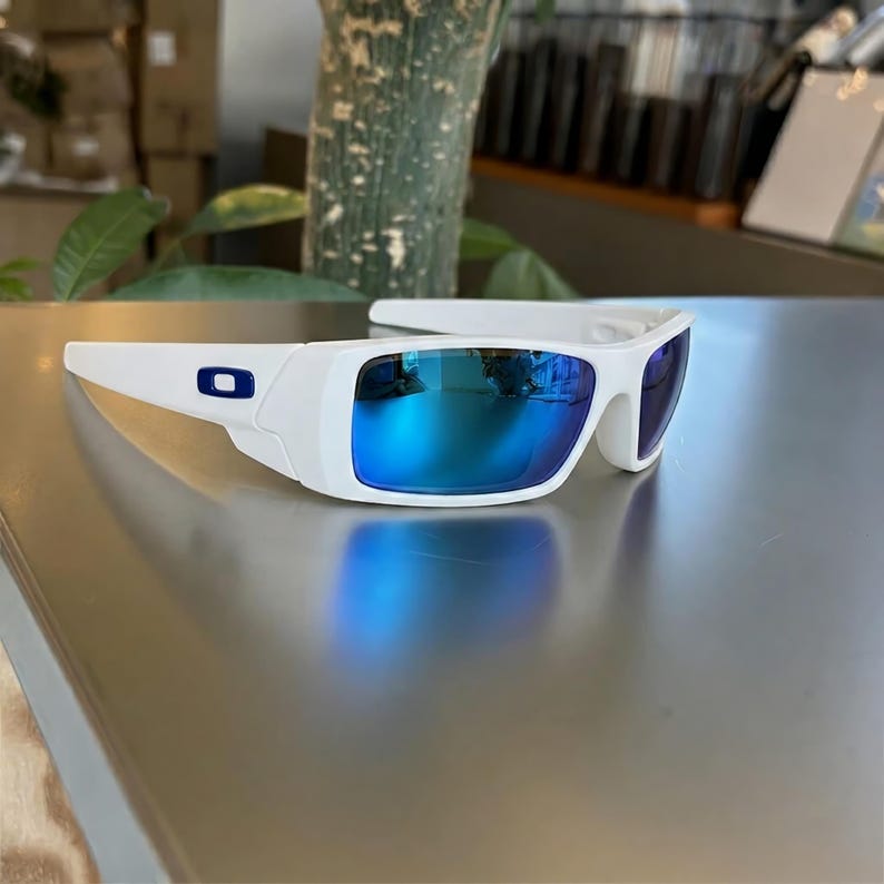May include: White sunglasses with blue mirrored lenses. The sunglasses have a rectangular frame with a blue logo on the side. The lenses reflect the surrounding environment. The sunglasses are on a reflective surface.